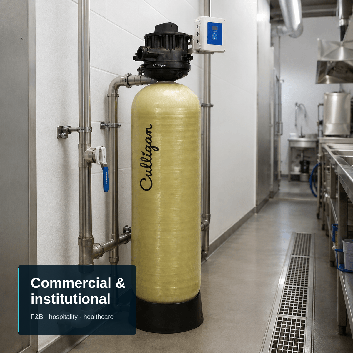 Editorial composition of a single Culligan CTM top-mount filter tank installed against the wall of a clean commercial utility corridor, connected to stainless-steel pipework with an isolation valve, with a floor drainage channel alongside and back-of-house equipment visible beyond. A compact text block anchored to the bottom-left carries the headline "Commercial & institutional" and the subtitle "F&B · hospitality · healthcare".